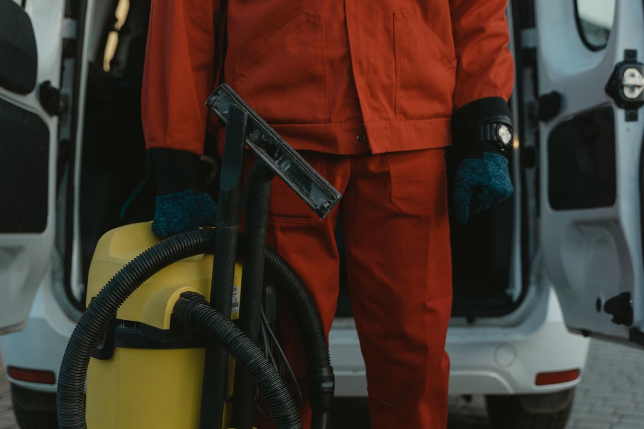 why-choose-us-02 A professional cleaner in a red uniform holding a vacuum near a van, ready for service.