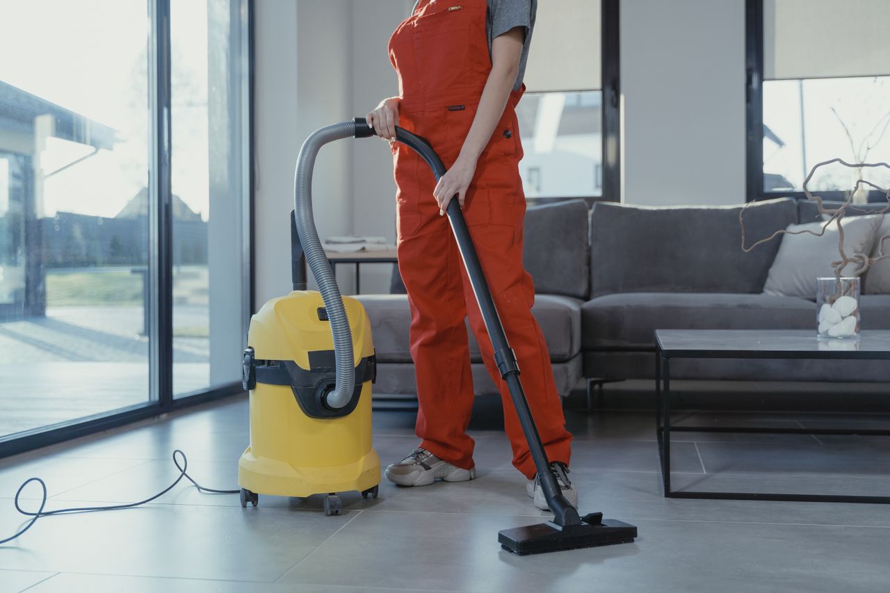 services-01 A professional cleaner in red coveralls vacuuming a modern living room with a yellow vacuum cleaner.